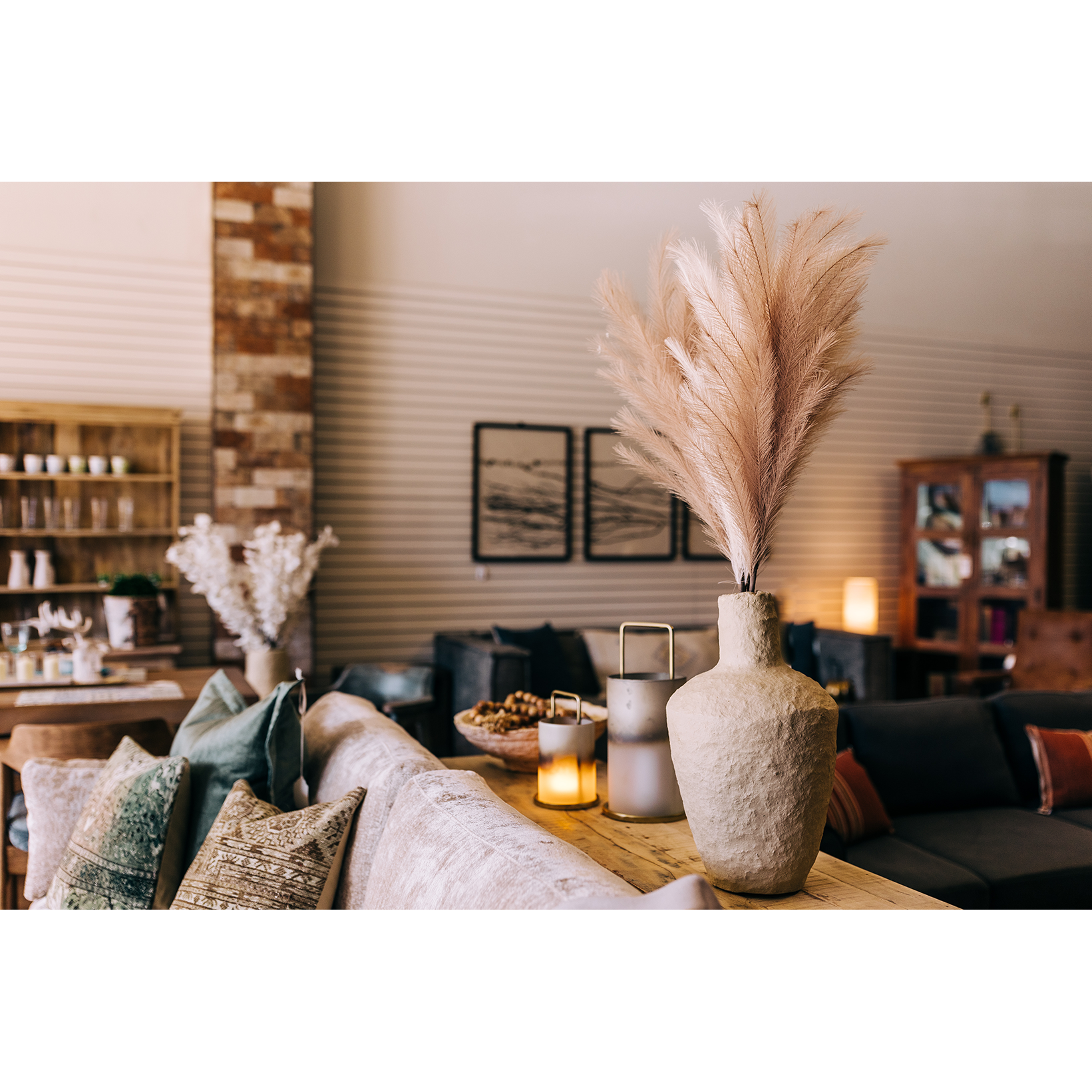 A cozy living room features a beige sofa with patterned pillows, a Paper Mache Vase filled with pampas grass on a wooden table, candles, and a decorative tray. Shelves with ceramics, framed art, and warm lighting make the space welcoming and stylish.