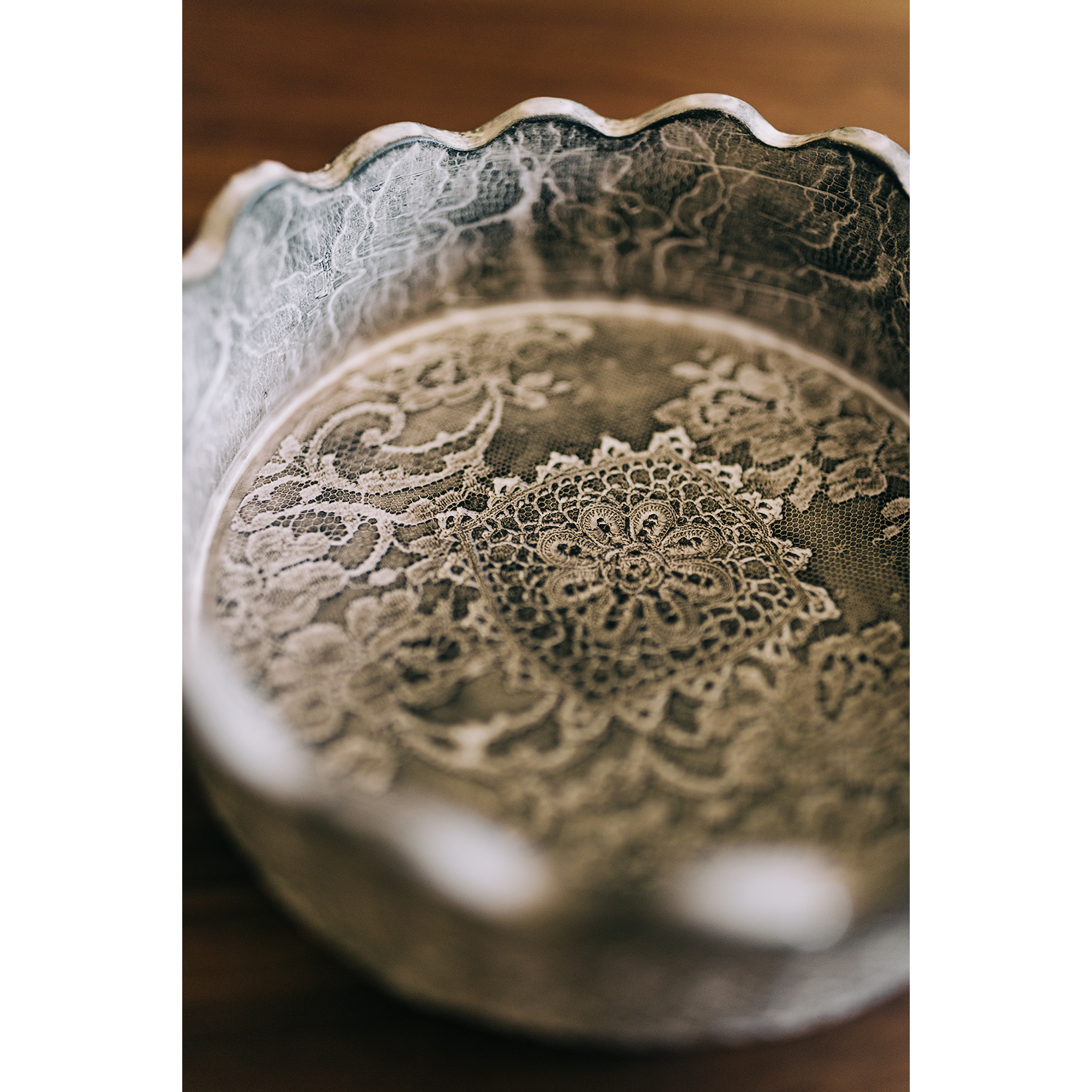 The Casado Crown Vessel is a round tray with scalloped edges, lined with vintage lace patterns of intricate floral and geometric designs. Placed on wood, it radiates an elegant, timeless charm.