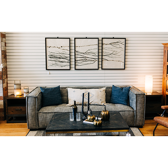 A modern living room showcases a Princeton Sofa in charcoal with tufted detailing, accented by blue and white cushions. A black coffee table holds decorative items like candles and gold pumpkins. Three black-and-white abstract artworks adorn the striped wall behind, while ambient lighting adds warmth.