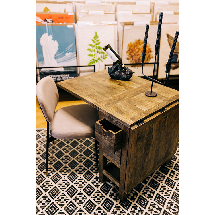A small wooden Harold Folding Table with storage slots on the side is placed on a black and white geometric patterned rug. An upholstered chair is pulled up to the desk. The table holds a black sculpture and a standing candle holder. Background shelves are filled with papers and artwork in various colors.