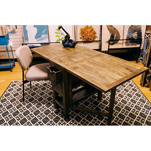 A Harold Folding Table with a black and white patterned rug underneath is paired with a beige cushioned chair. On the table, there's a sculpture of a black horse and a small plant with green leaves. The background features various decorative panels and items displayed on shelves, perfect for a small space.
