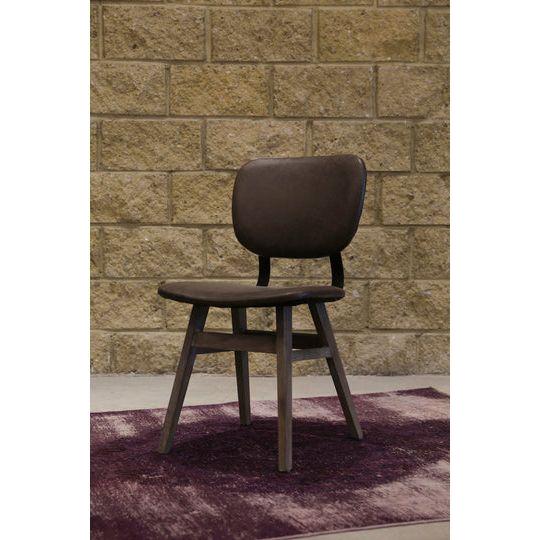 A Brown Leather Dining Chair made of reclaimed wood with a cushioned backrest and brown leather seat is positioned on a dark purple rug. The background features a textured brick wall in shades of tan and beige. The chair has four legs and a simple, modern design.