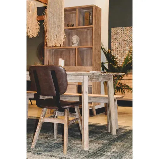 A rustic dining area features a wooden table and chairs. The table is rectangular with a natural, unfinished wood look, complemented by Brown Leather Dining Chairs with reclaimed wood bases. Behind the table is a tall bookshelf adorned with various decorations and plants in the background.