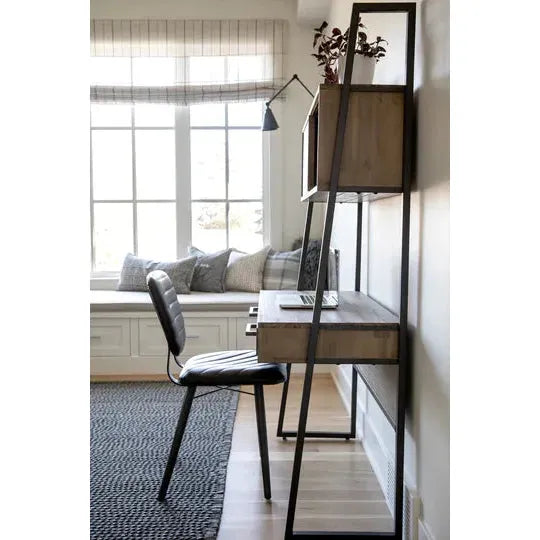 A modern home office space features a minimalist desk with a black metal frame and light wood surfaces. On the desk is a closed laptop. Above it, a shelf holds a small plant. A luxurious Black Dining Chair faces the desk, and a nearby window with a window seat has cushions and a plaid valance.