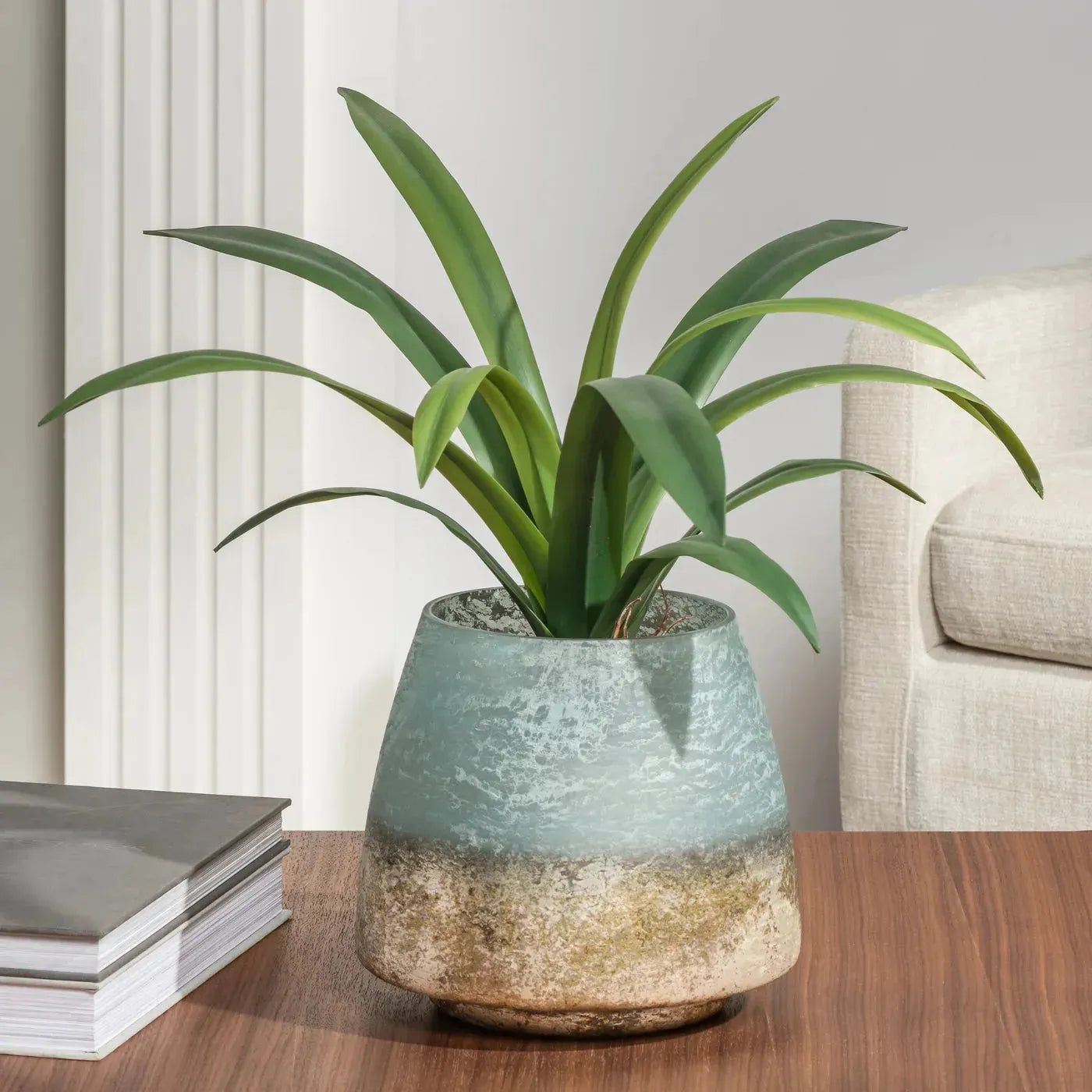 A green, broad-leaved plant is elegantly displayed in a Cambridge Hurricane Vase that features a rustic ombre design transitioning from brown at the base to blue at the top. This versatile home decor piece adorns a wooden table adjacent to a neatly stacked pair of books. In the background, part of a white column and a beige armchair can be seen.