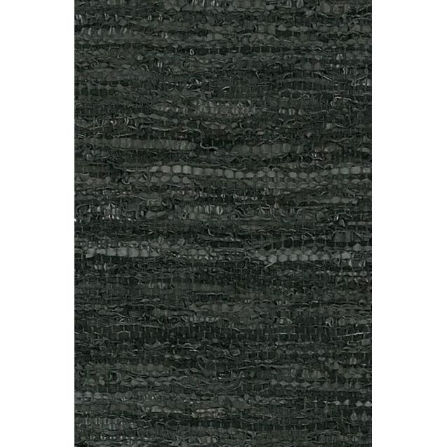 A close-up view of the Saket Rug reveals a dark textured fabric with a predominantly black color. The rug features a woven pattern made up of various shades of gray and black threads, resulting in an irregular, wavy appearance. Its texture consists of a blend of thicker and thinner strands, giving it a slightly rough surface.