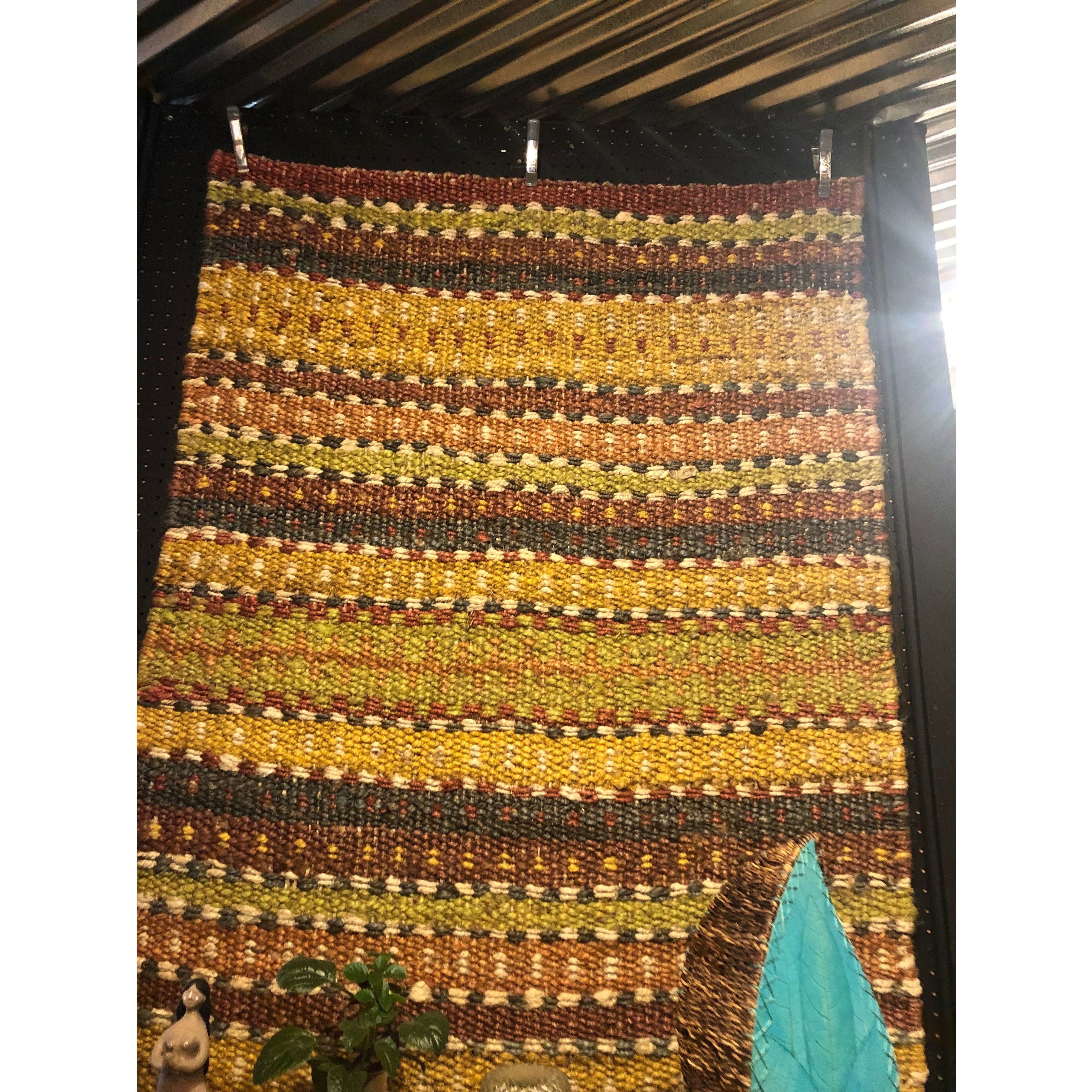 The Saket Rug, a colorful woven tapestry featuring horizontal stripes in shades of brown, yellow, green, and white, hangs on a vertical surface. At the bottom of the rug, a blue fabric decoration and some plants are partially visible. The rug is secured with metal clips at the top.