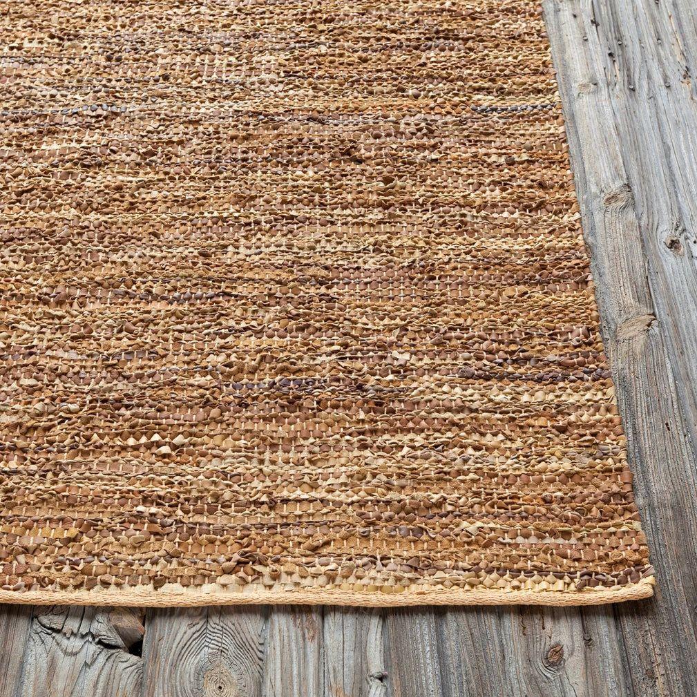 A close-up image showcases the Saket Rug, featuring a woven design in varying shades of beige and light brown. The textured area rug is placed on a wooden floor, highlighting its grain and knots. The combination of the Saket Rug's intricate weave and the rustic wooden floor creates a warm, earthy ambiance.