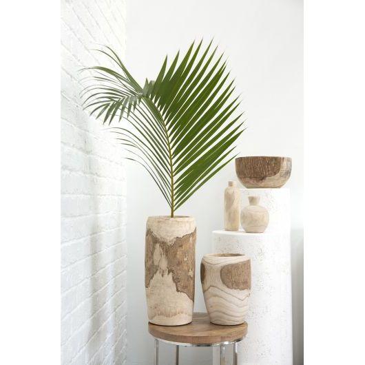A minimalist arrangement features a tall, cylindrical Yucca Vase with a single green palm frond. The handcrafted vase is placed on a small round wooden table. In the background, there are various wooden vases and bowls arranged on a white pedestal against a white brick wall, embodying contemporary design.