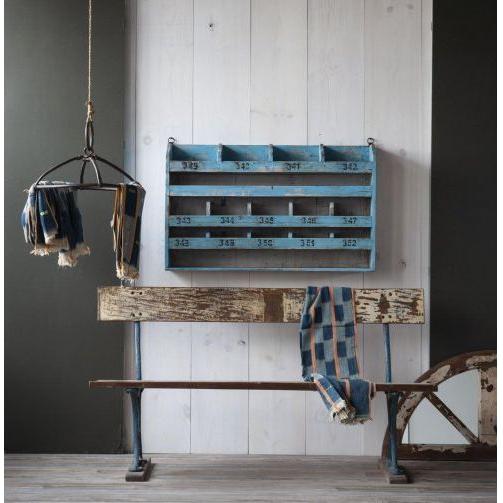 A rustic room features a green and white paneled wall adorned with a weathered wooden bench. Adding character, the Cubby Hole Hanging Shelf with 40'' wide numbered cubbies is mounted above. A hanging light fixture displays fabric pieces, while a plaid scarf is draped over the bench, and a wooden wheel peeks out on the right.