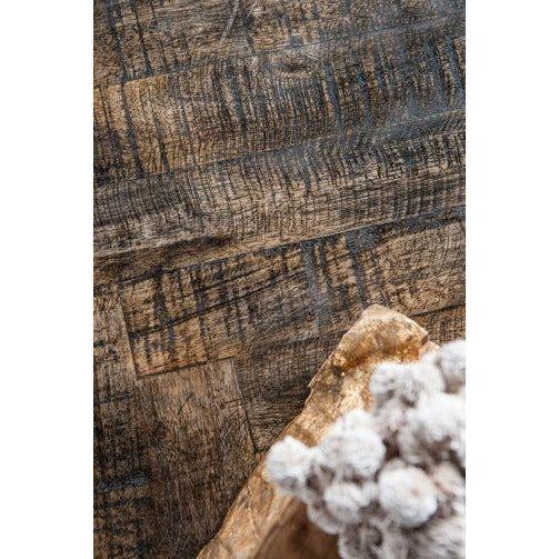 A close-up of a rustic wooden floor showcases a rich, textured grain pattern in shades of brown and grey. The Parquette Coffee Table, featuring a brass finish, enhances the cozy atmosphere, while beige fabric in the foreground adds an element of softness. The combination of textures and finishes crafts a warm setting.