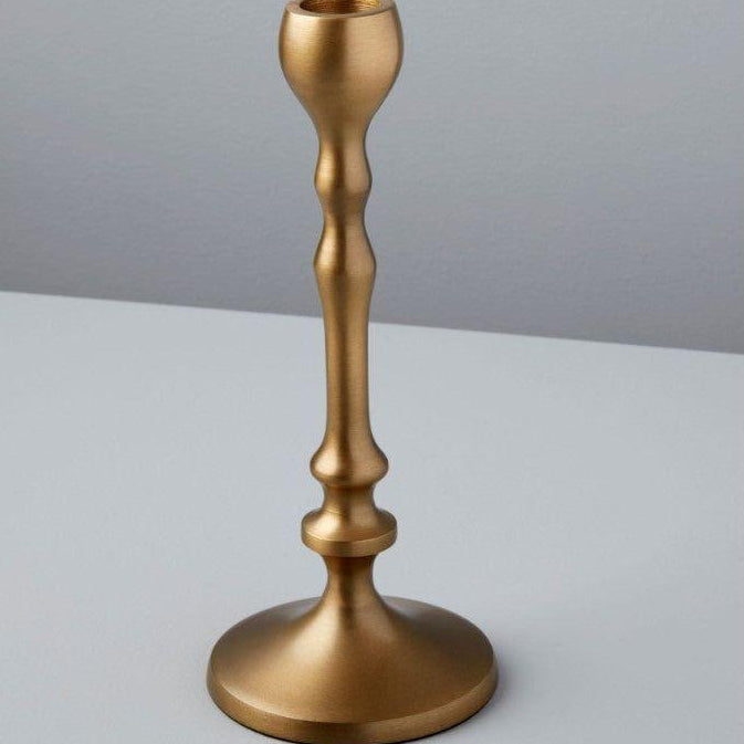 An aged bronze candlestick stands on a light gray surface against a pale gray background. This handcrafted piece features a smooth, curved design with a wide circular base and narrow top, perfect for holding one candle. Its metallic finish exudes the elegance of luxurious home essentials.