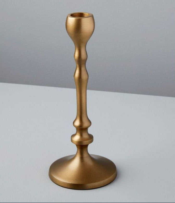 An aged bronze candlestick stands on a light gray surface against a pale gray background. This handcrafted piece features a smooth, curved design with a wide circular base and narrow top, perfect for holding one candle. Its metallic finish exudes the elegance of luxurious home essentials.