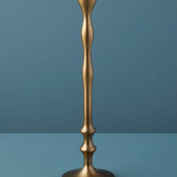 A sleek, modern candlestick holder, reminiscent of the Aged Bronze Candlestick, stands on a flat surface. This handcrafted piece boasts a gracefully sculpted, undulating column and a round base. Set against a solid teal backdrop, its elegant metallic sheen epitomizes luxurious home essentials.