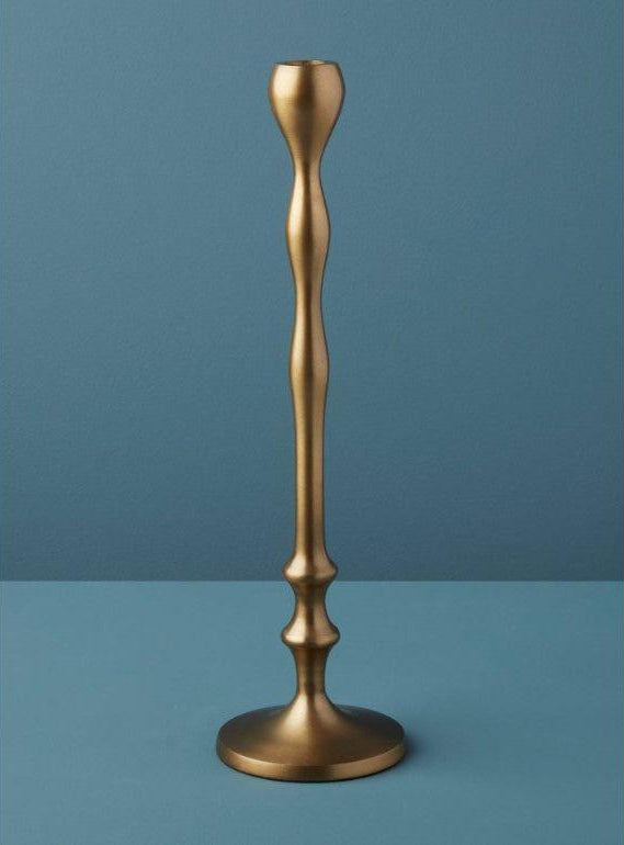 A sleek, modern candlestick holder, reminiscent of the Aged Bronze Candlestick, stands on a flat surface. This handcrafted piece boasts a gracefully sculpted, undulating column and a round base. Set against a solid teal backdrop, its elegant metallic sheen epitomizes luxurious home essentials.