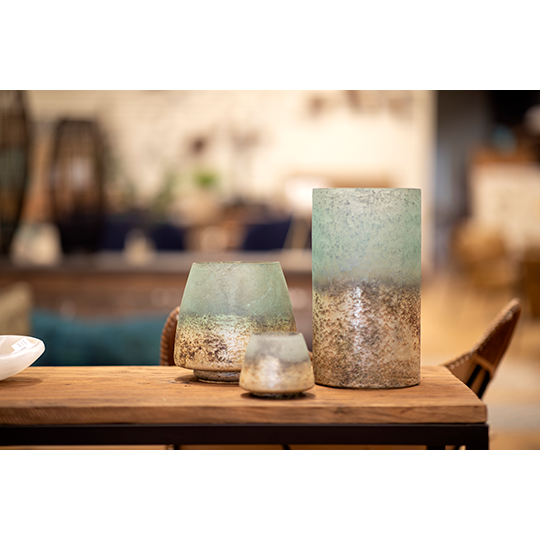 Three Cambridge Hurricane Vases of varying sizes sit on a wooden table. Each vase features a greenish hue transitioning to an antiqued gold base with a rustic texture. The background appears to be a cozy, warmly lit room with blurred furniture and decor elements, perfect for versatile home décor.