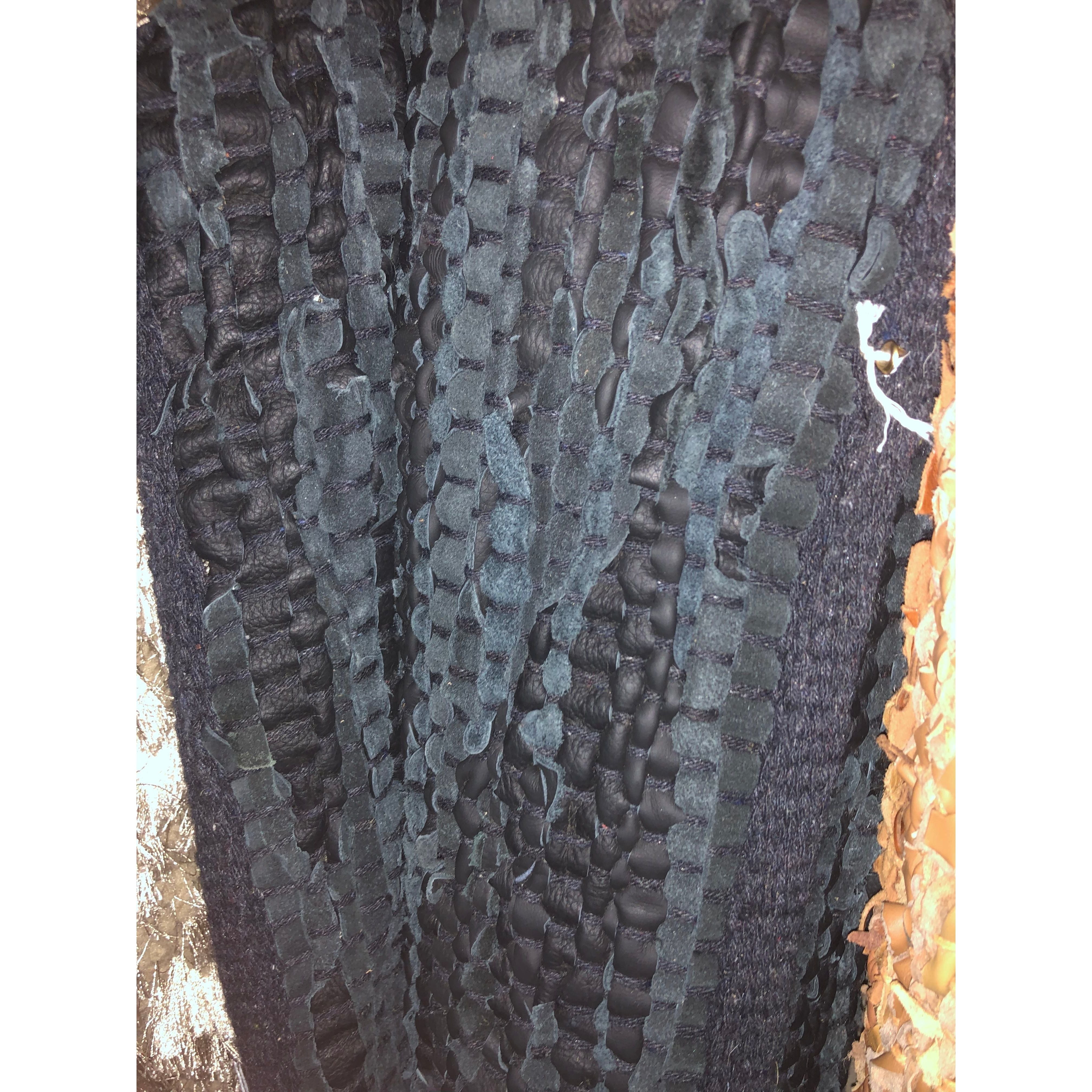 A close-up of the Saket Rug showcases its textured fabric, featuring a pattern of thick, intertwining black and dark grey woven strips. The rugged, uneven surface creates a tactile, layered appearance. To the right, partially visible, is a contrasting section in beige and tan hues.
