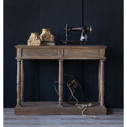 A rustic table with a Carved Pillar Base in natural wood holds an antique sewing machine and two ornate gold-toned jars. A cord is coiled on the lower shelf. The dark minimalist wall contrasts with the light-colored wood floor.