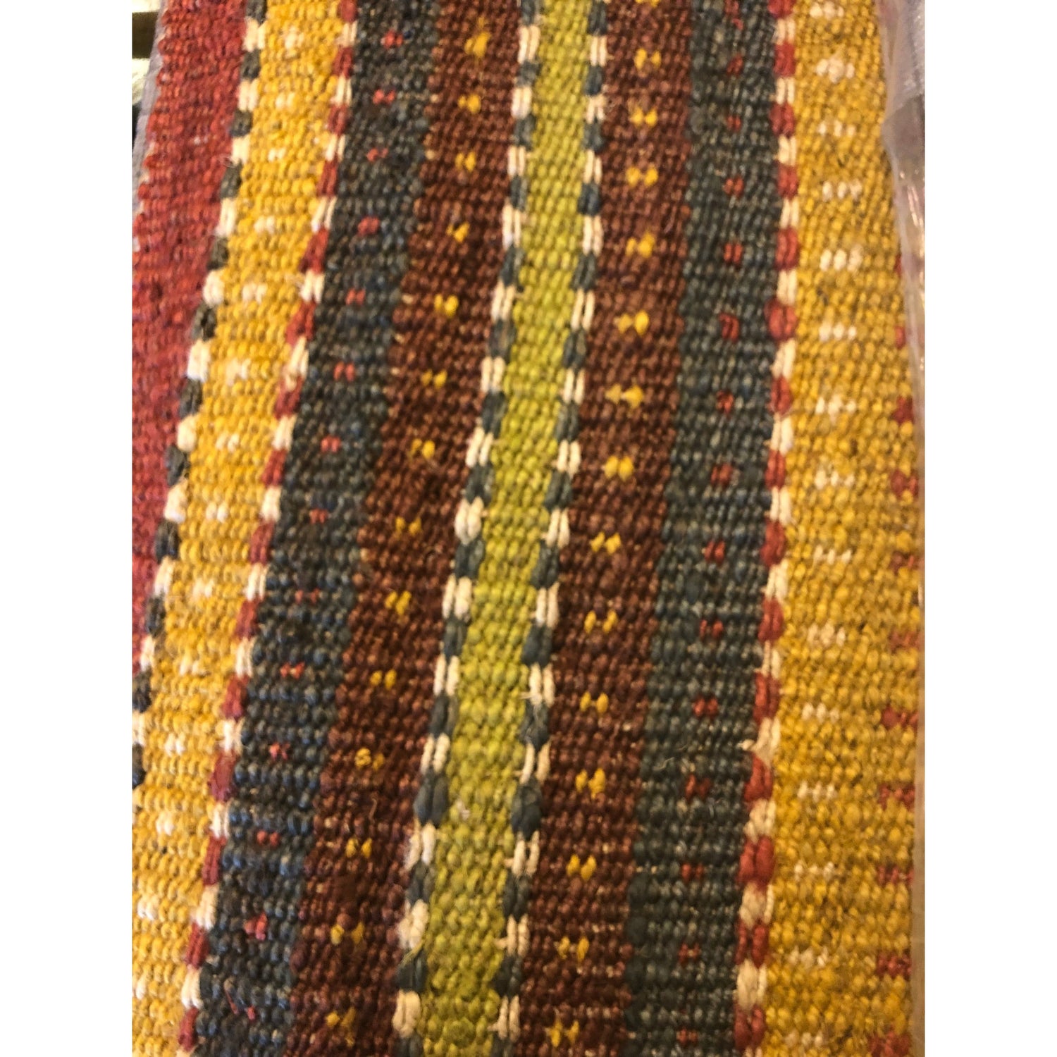 A close-up of the Saket Rug reveals its woven textile featuring vertical stripes in yellow, brown, green, and red. The stripes display uneven, dotted patterns in contrasting colors, complemented by smaller white and blue accents. The texture appears coarse and handmade, with visible threads that contribute to its rustic aesthetic.
