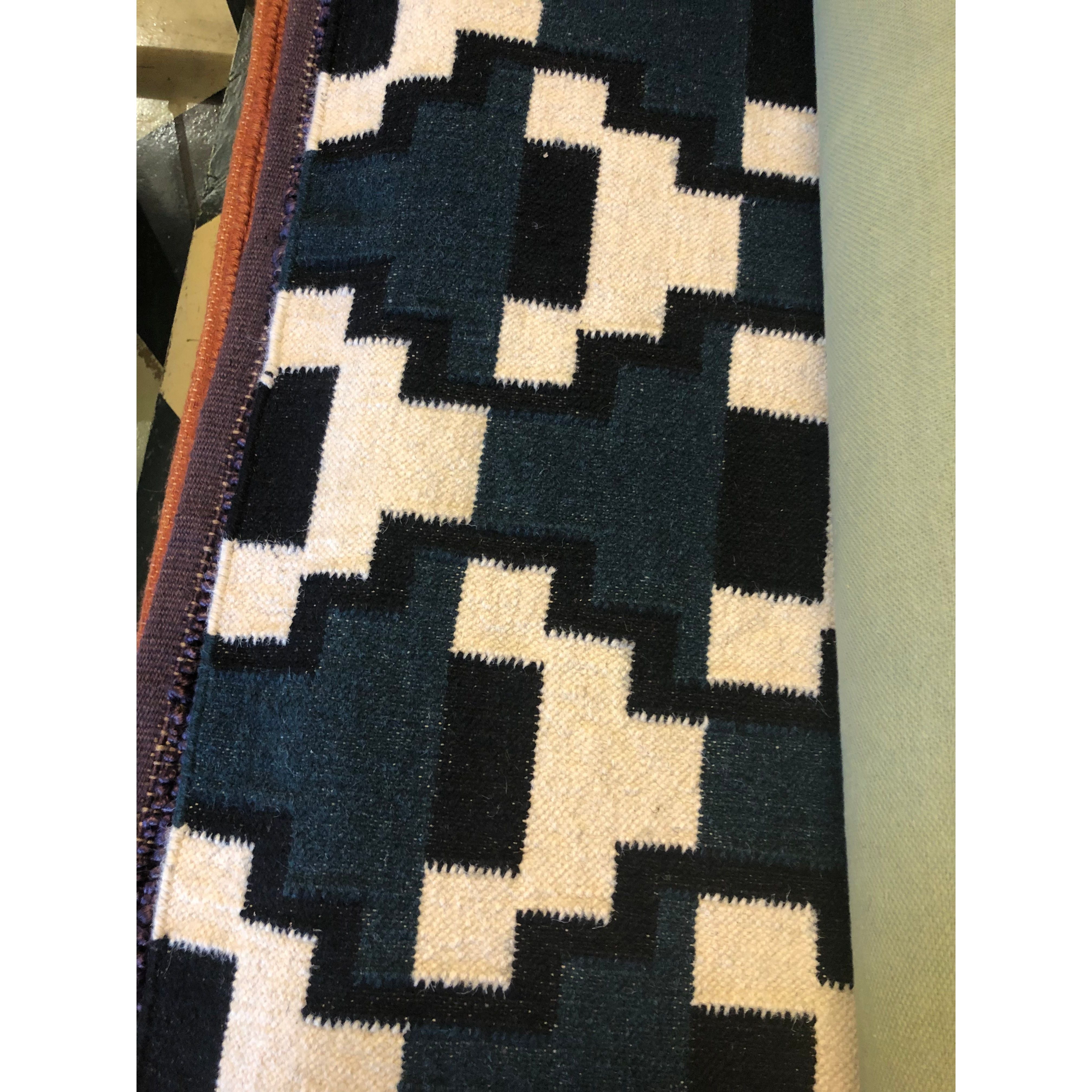 A close-up of the Avon Wool Rug shows cream and dark green zigzag rectangles in a bold, repeating geometric pattern. The rug is rolled, with its edges and some nearby materials also visible.