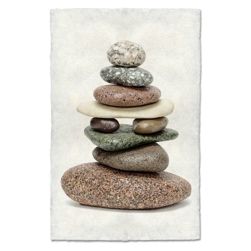 Balanced (1) features a vertical stack of seven smooth, earth-toned stones, arranged largest to smallest, on textured white handmade paper that evokes Nepalese craftsmanship and individuality.
