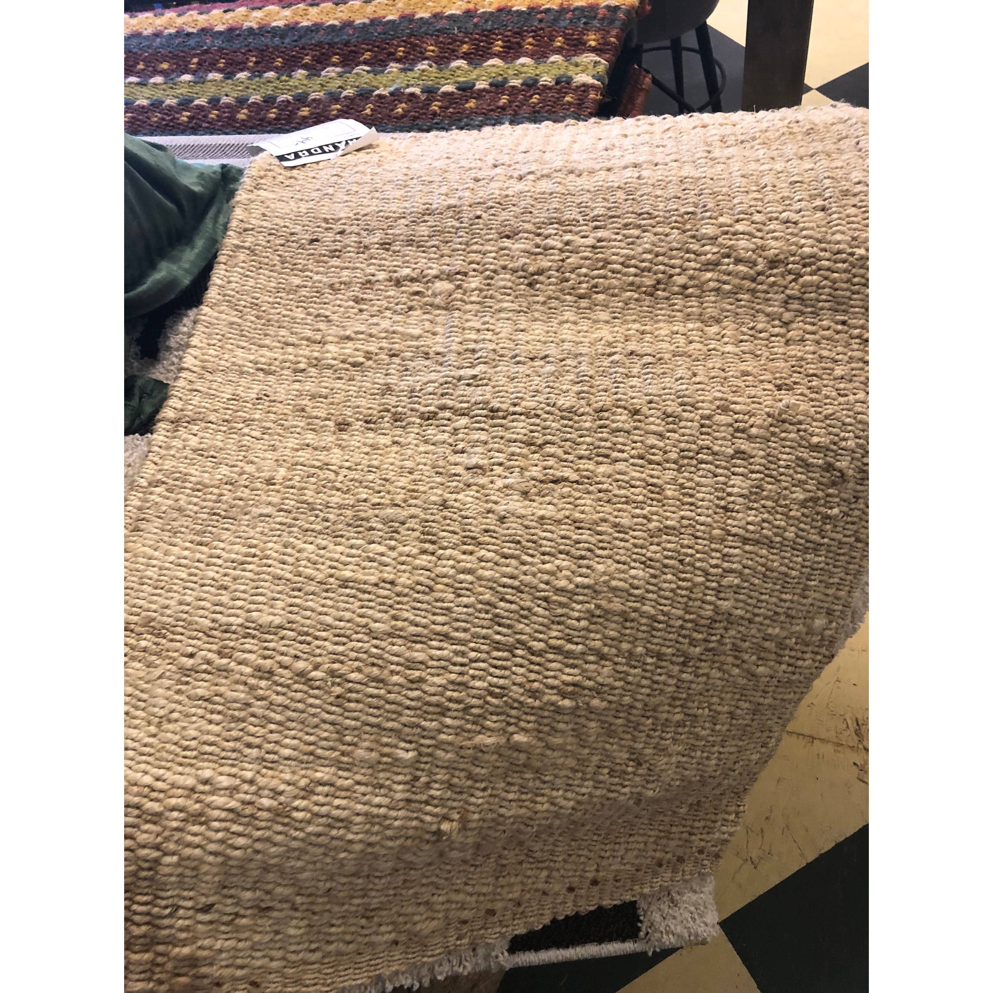 A Saket Rug in textured beige is draped over a dark wooden table, showcasing its woven, chunky appearance with uneven yarn thickness. In the background, an orange and brown patterned rug is partially visible on what appears to be a checkered black-and-white tile floor.