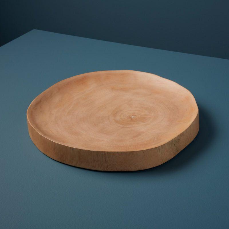 A smooth, round Wood Charger crafted from beautifully handcrafted mango wood rests on a blue surface. The charger showcases a light, natural color with slightly curved edges. The darker blue background adds contrast to this sustainably sourced piece, creating an overall minimalist and elegant effect.