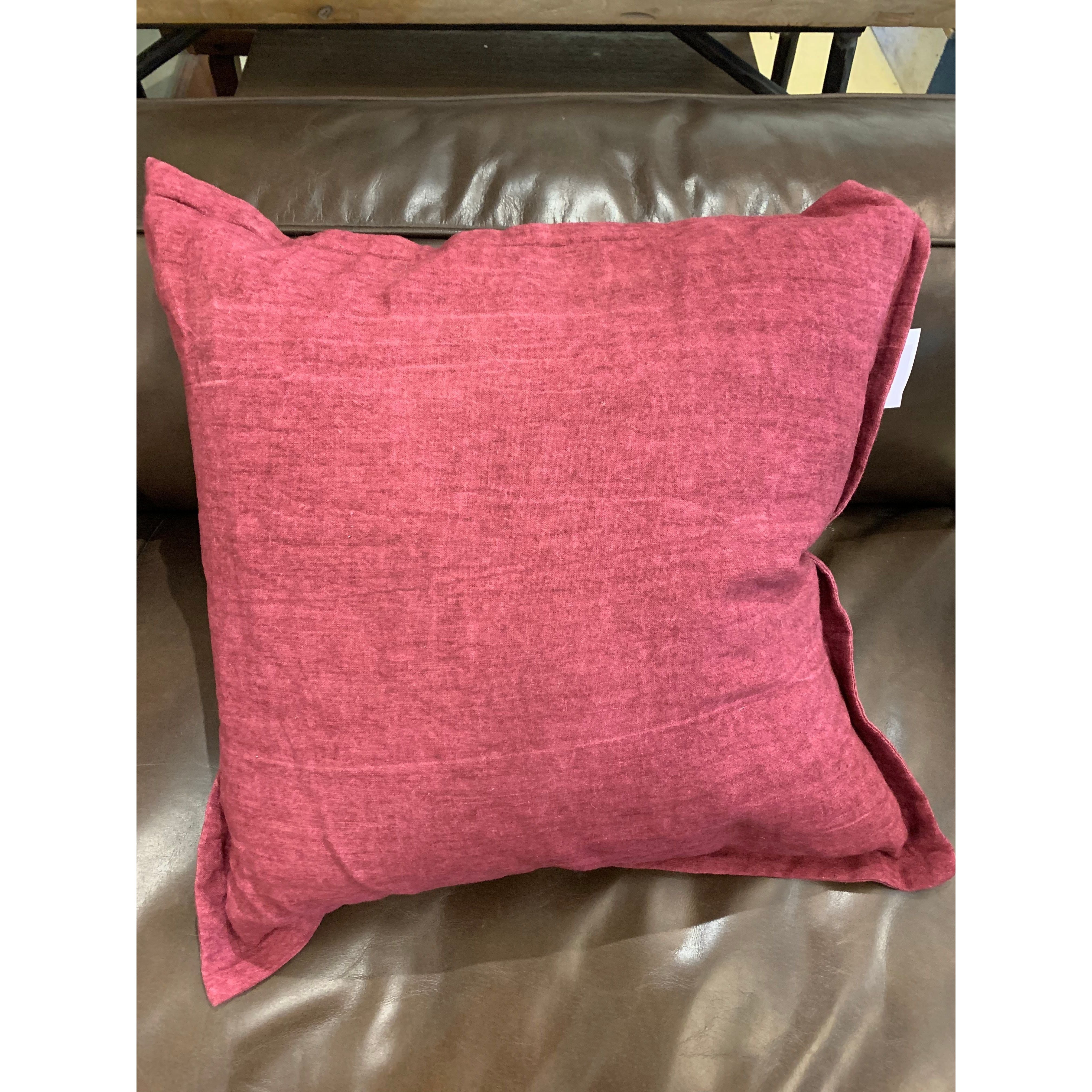 A square, red Throw Pillow made of fabric with a distressed effect rests on a brown leather couch. The slightly worn edges and bent corners contribute to the casual vibe of the setup. A white tag stands out on the top right side of the pillow, contrasting its subtly textured look.