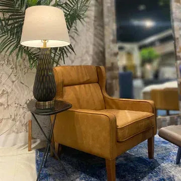 An Italian Leather Accent Chair with wooden legs sits on a blue patterned rug beside a small black tripod-base side table topped with a dark textured lamp and white shade, with tall green palm leaves in the background.