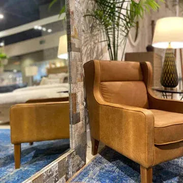 An Italian Leather Accent Chair with wooden legs sits on a blue rug beside a tall mirror. Behind it are a decorative lamp with a textured base and a leafy green plant, both reflected in the mirror against a modern backdrop.