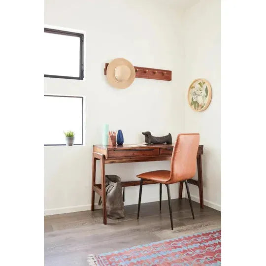 A small home office includes a wooden desk paired with the Lucy Leather Dining Chair in tan. Books, vases, and a seal-shaped sculpture sit on the desk. A straw hat hangs on a black iron wall rack above. A potted plant adorns the window ledge to the left, over a patterned rug on the wooden floor.