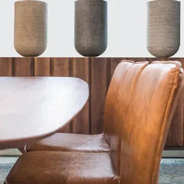 A modern dining area features a wooden table flanked by Lucy Leather Dining Chairs in tan. A black iron frame highlights the wooden-paneled wall behind the table, which is adorned with three cylindrical gray planters above, adding a minimalist decor touch.