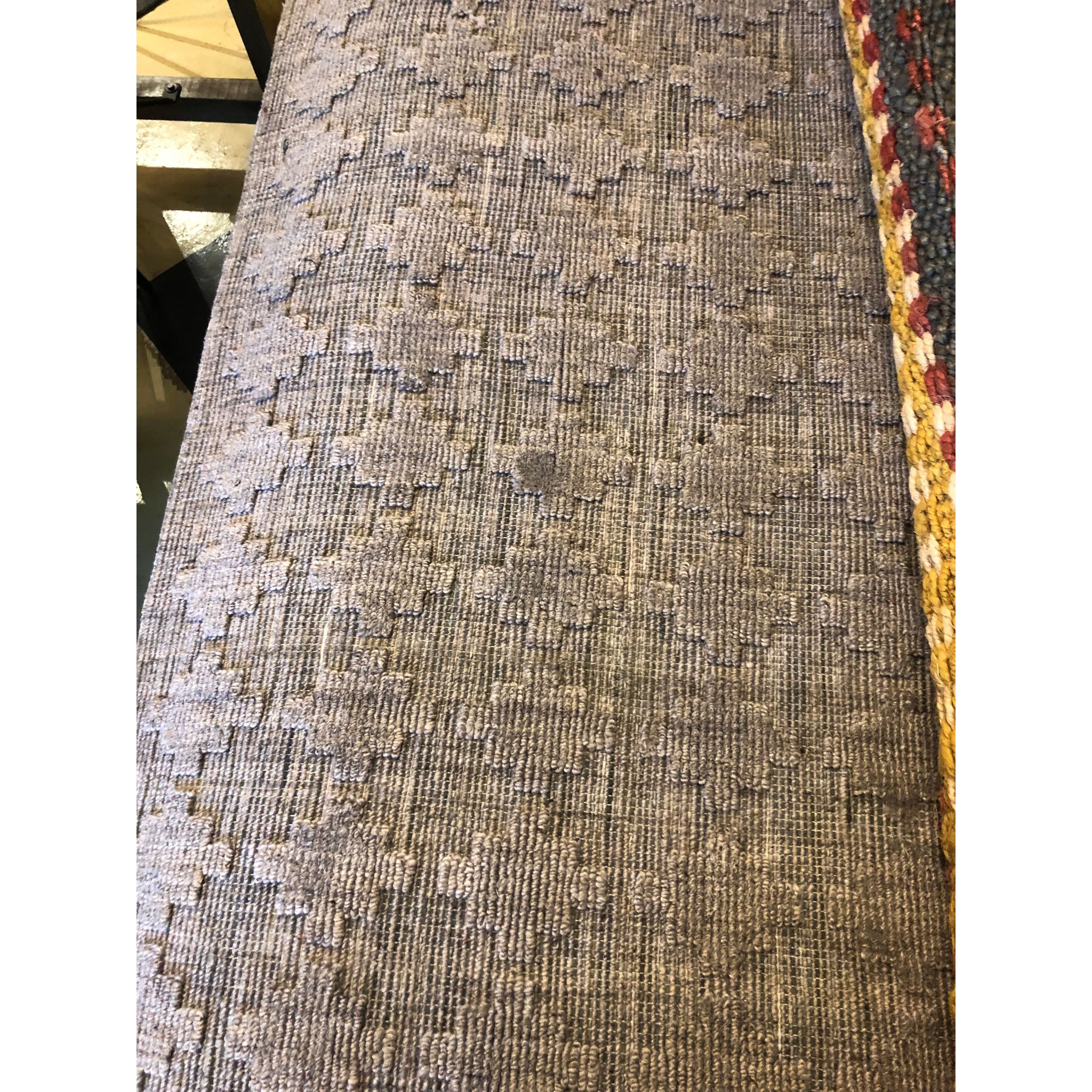 Close-up of the Netix Wool/Viscose Rug featuring a textured surface with a subtle geometric, tile-like pattern in muted purple-gray hues and a thick woven trim in red, yellow, and brown along one edge.