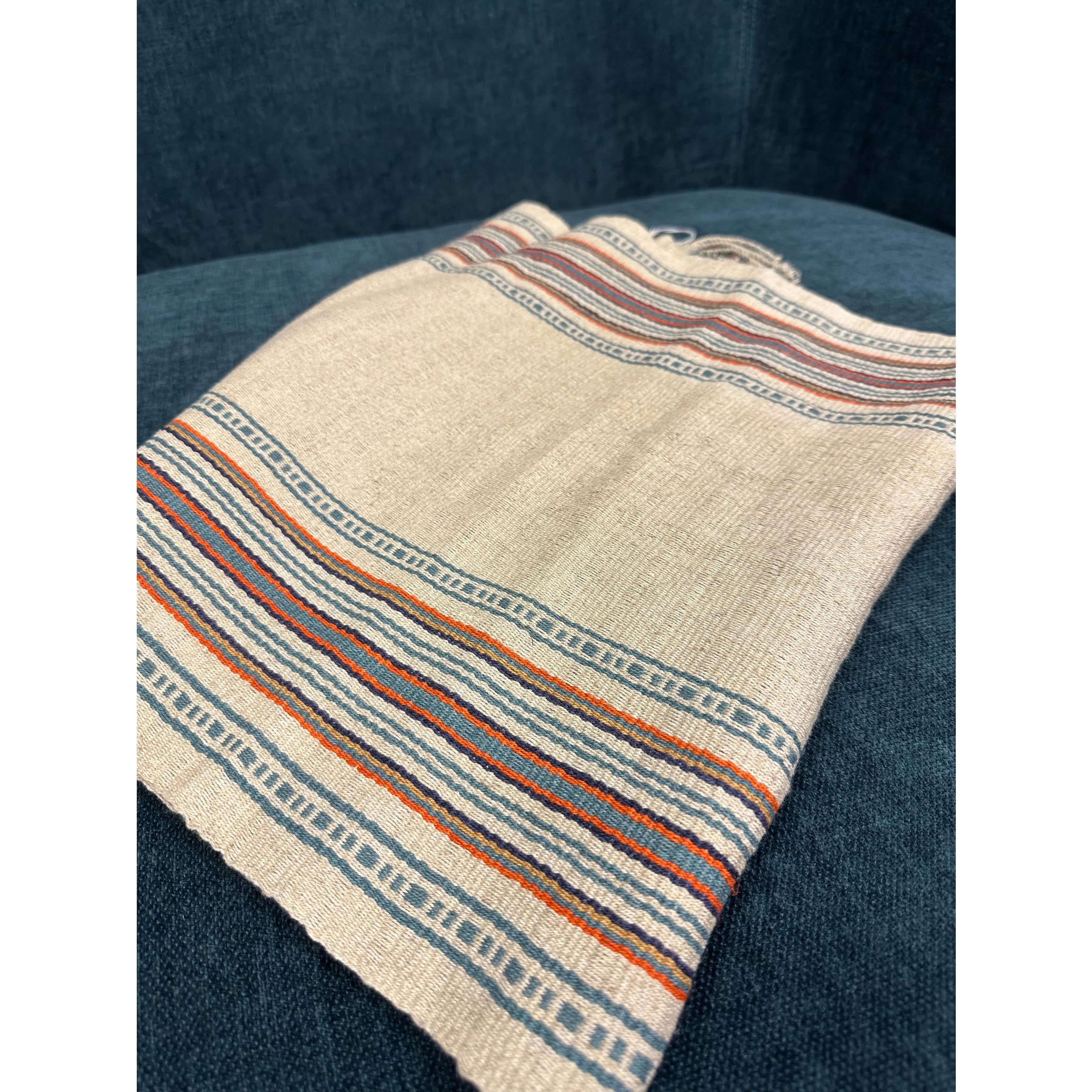 A close-up of the Hemp Table Runner featuring a beige woven fabric adorned with horizontal striped patterns in muted red, blue, and navy blue. This handwoven table runner, crafted by skilled artisans, showcases intricate geometric designs and is displayed on a dark blue upholstered surface.