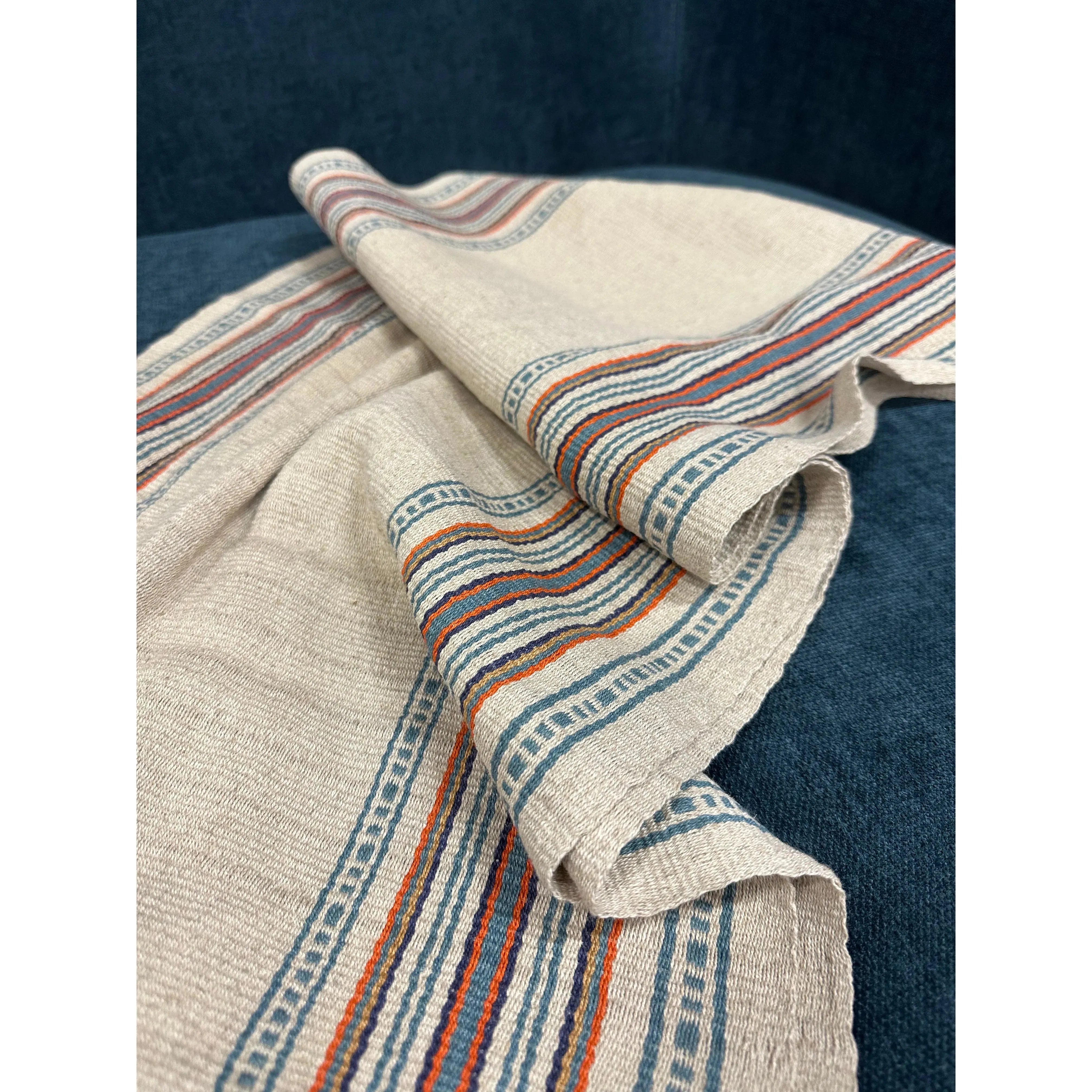A close-up of the Hemp Table Runner in beige, adorned with intricate borders in shades of blue, orange, and red. Handwoven by skilled artisans, it rests on a dark blue fabric background. The slightly crumpled appearance highlights the texture of the fabric and the detailed woven designs along its edges.