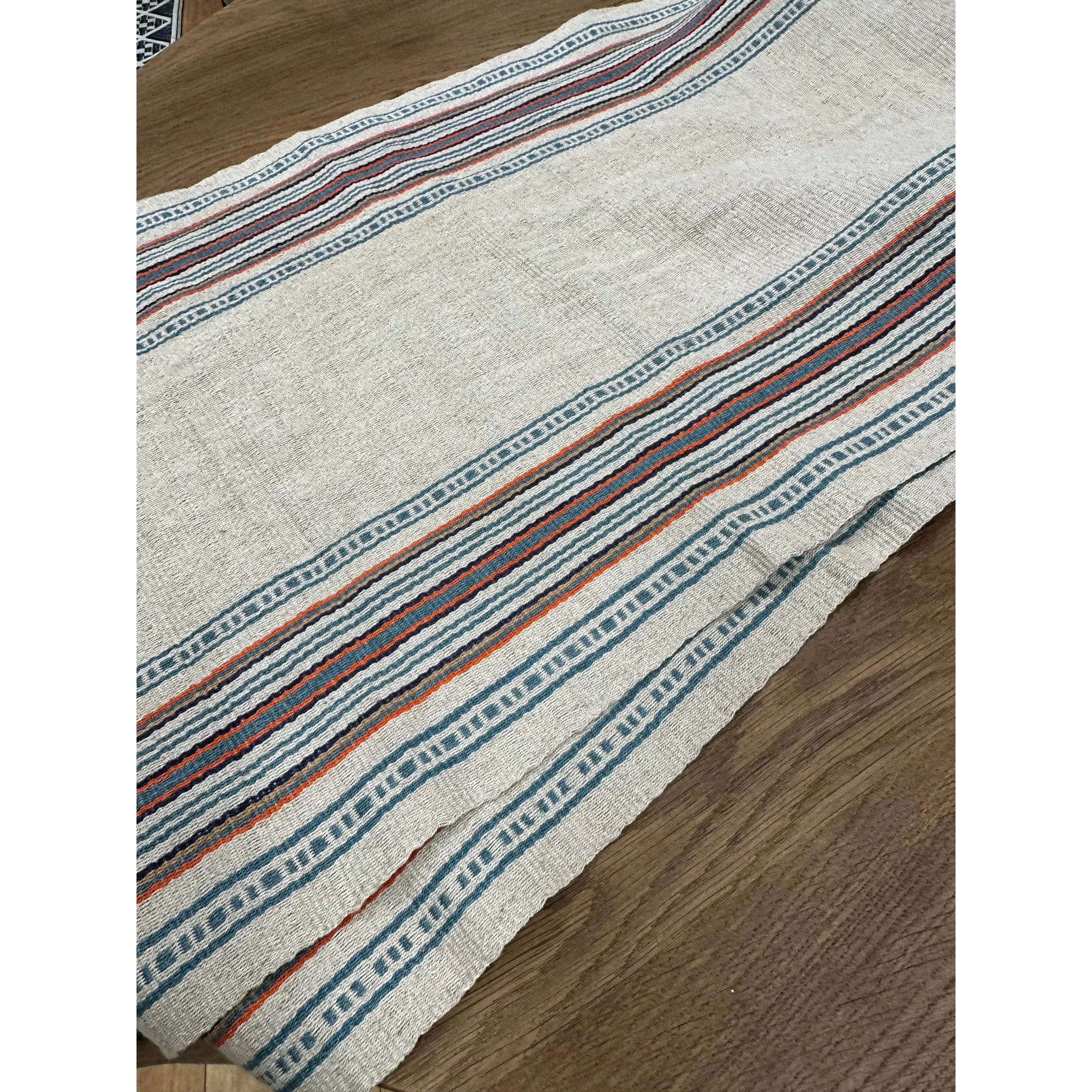 A close-up of the Hemp Table Runner, skillfully handwoven and laid out on a wooden table. This runner boasts a beige background adorned with decorative horizontal stripes in blue, red, and orange. The blue stripes showcase intricate, repeating geometric patterns, while the red and orange stripes are solid lines meticulously crafted by skilled artisans.