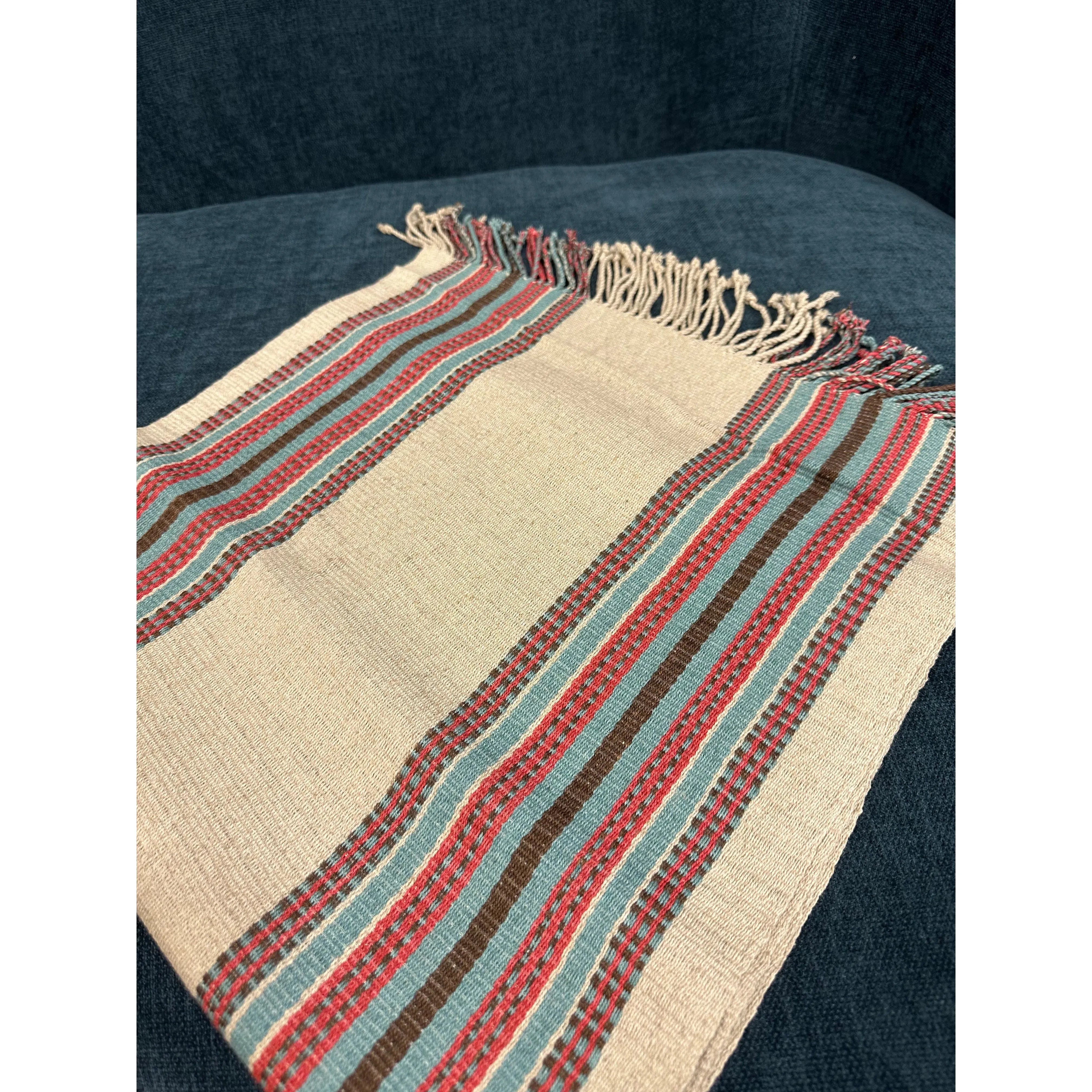 The Hemp Table Runner, handwoven by skilled artisans, showcases horizontal stripes in a variety of colors such as red, green, brown, and black. It is set against a dark blue fabric background and features short fringe tassels along its edges. The texture of the table runner is coarse and slightly uneven.