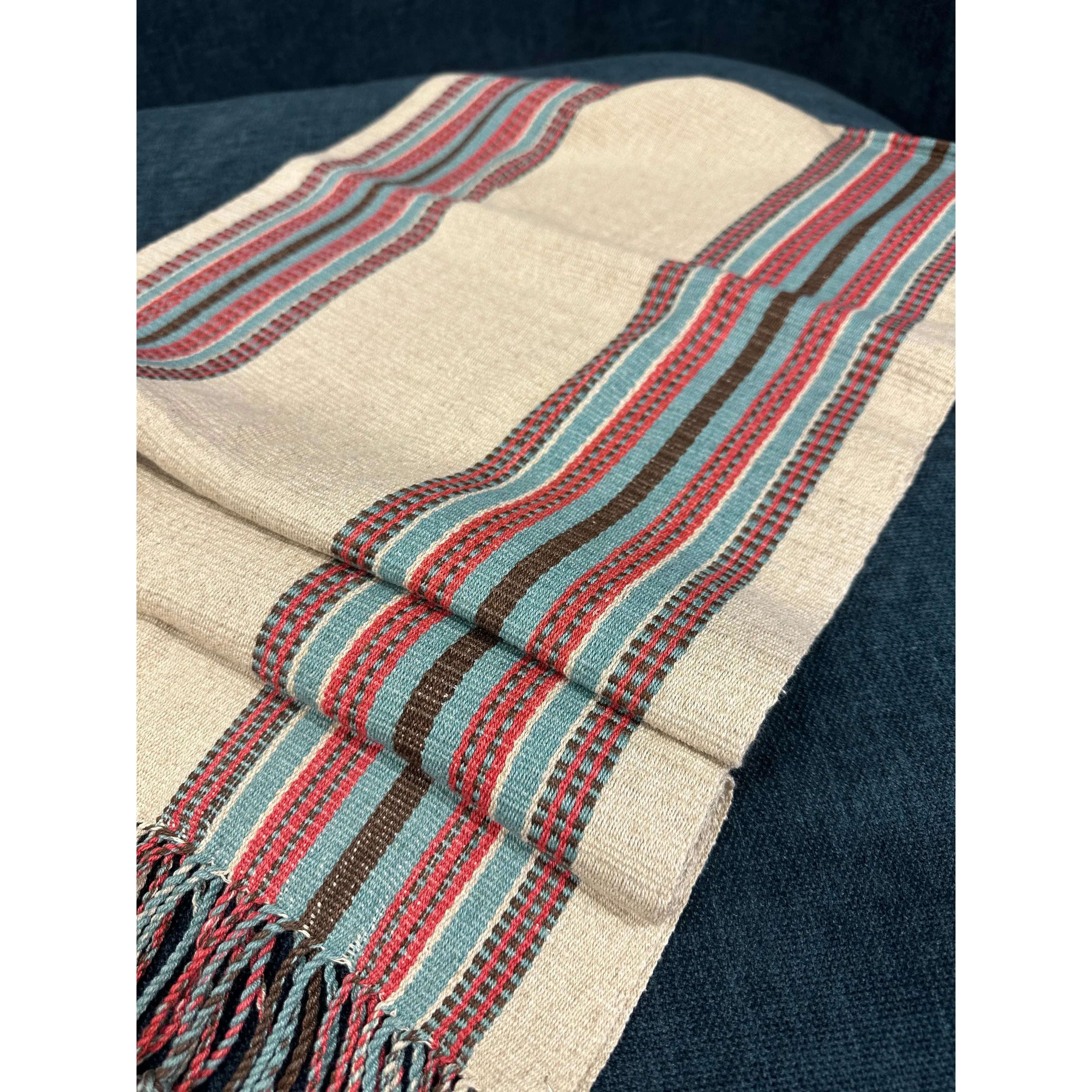 A Hemp Table Runner is draped over a dark surface. This beige, handwoven textile features vertical stripes in red, green, and brown, with plaid-like patterns interspersed throughout the bands. The table runner ends in a short fringe, showcasing the intricate patterns and colors meticulously crafted by skilled artisans.