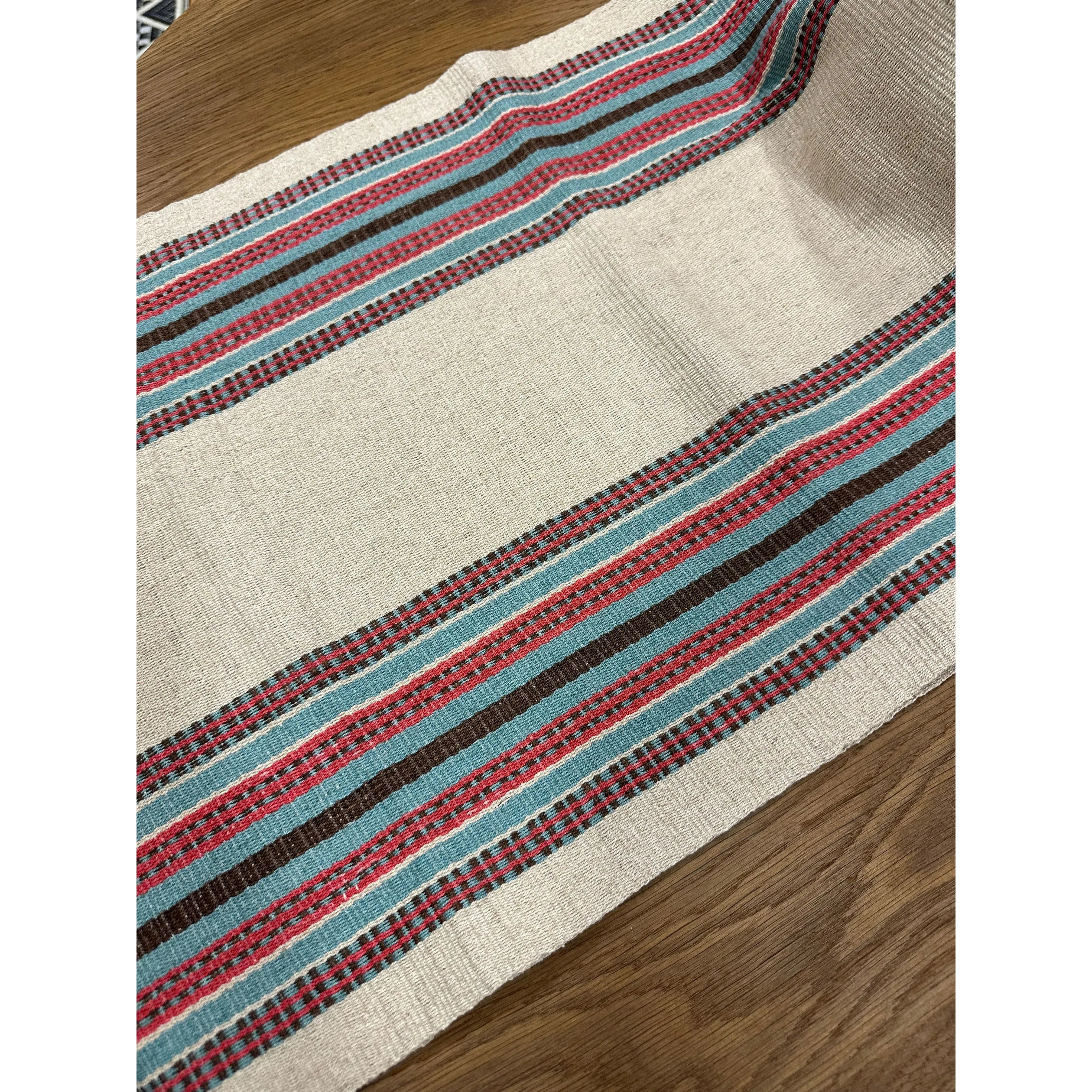A close-up image of the Hemp Table Runner on a wooden surface. The runner showcases stripes in red, blue, and black against a beige background. Crafted by skilled artisans, the pattern consists of horizontal lines, some narrow and some broad, creating a decorative, rustic design.
