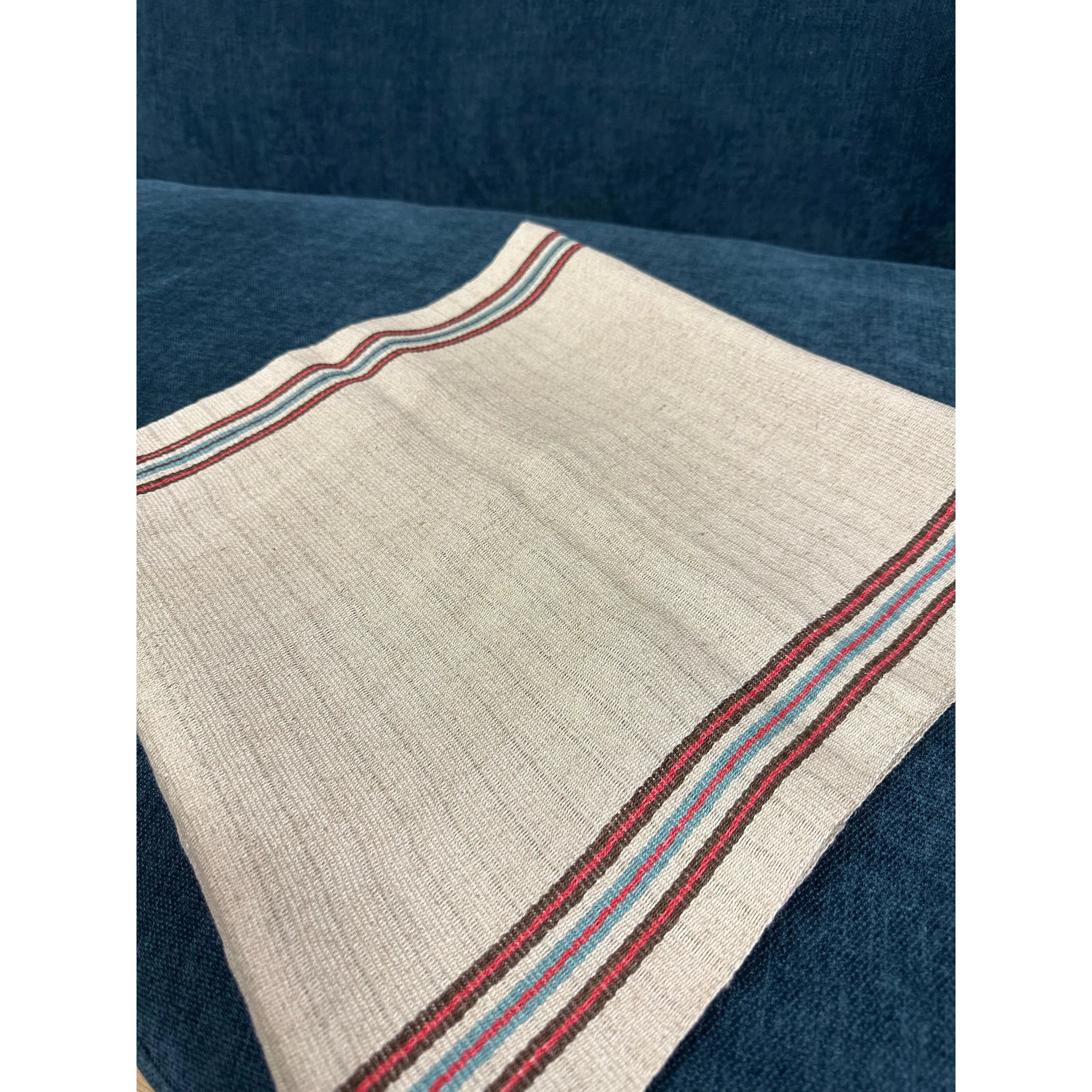 A Hemp Table Runner, composed of a square piece of beige fabric with multicolored stripes along two edges, is displayed on a dark blue surface. Handwoven by skilled artisans, the vibrant stripes in red, blue, and other colors offer a striking contrast to the plain background of the table runner. The texture showcases visible ribbed lines.
