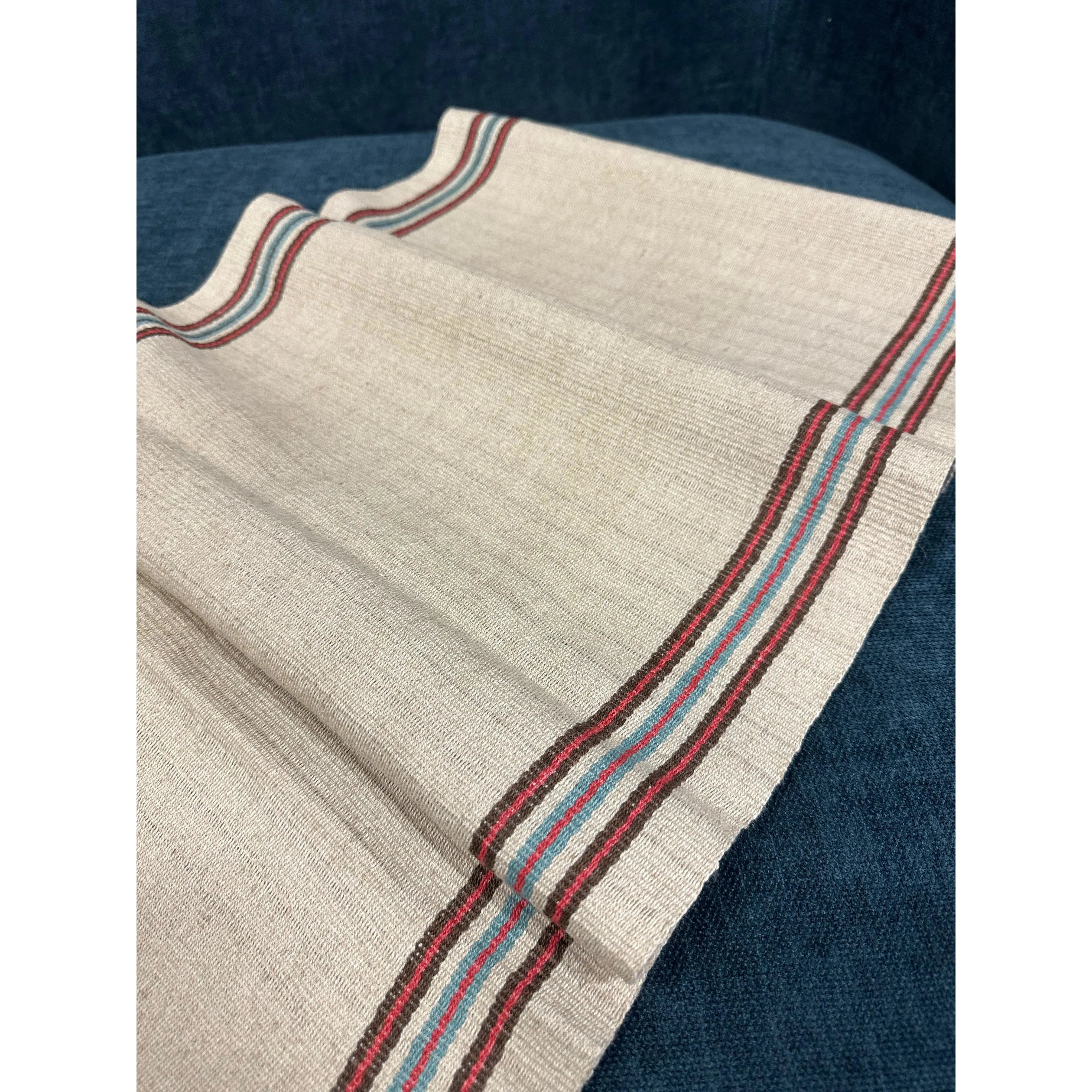 A beige textile cloth featuring fine horizontal texture patterns is elegantly presented against a dark blue fabric backdrop. The Hemp Table Runner, handwoven by skilled artisans, showcases colorful stripes in red, blue, and teal along its edges, providing a decorative border to the otherwise simple fabric.