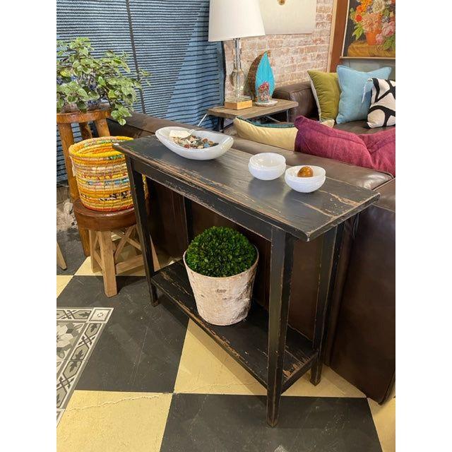A Reba Console Table (rustic black, reclaimed pine) stands behind a brown sofa, topped with ceramic dishes. Underneath, a wicker basket and potted plant add eco-friendly charm and rustic elegance to the room.
