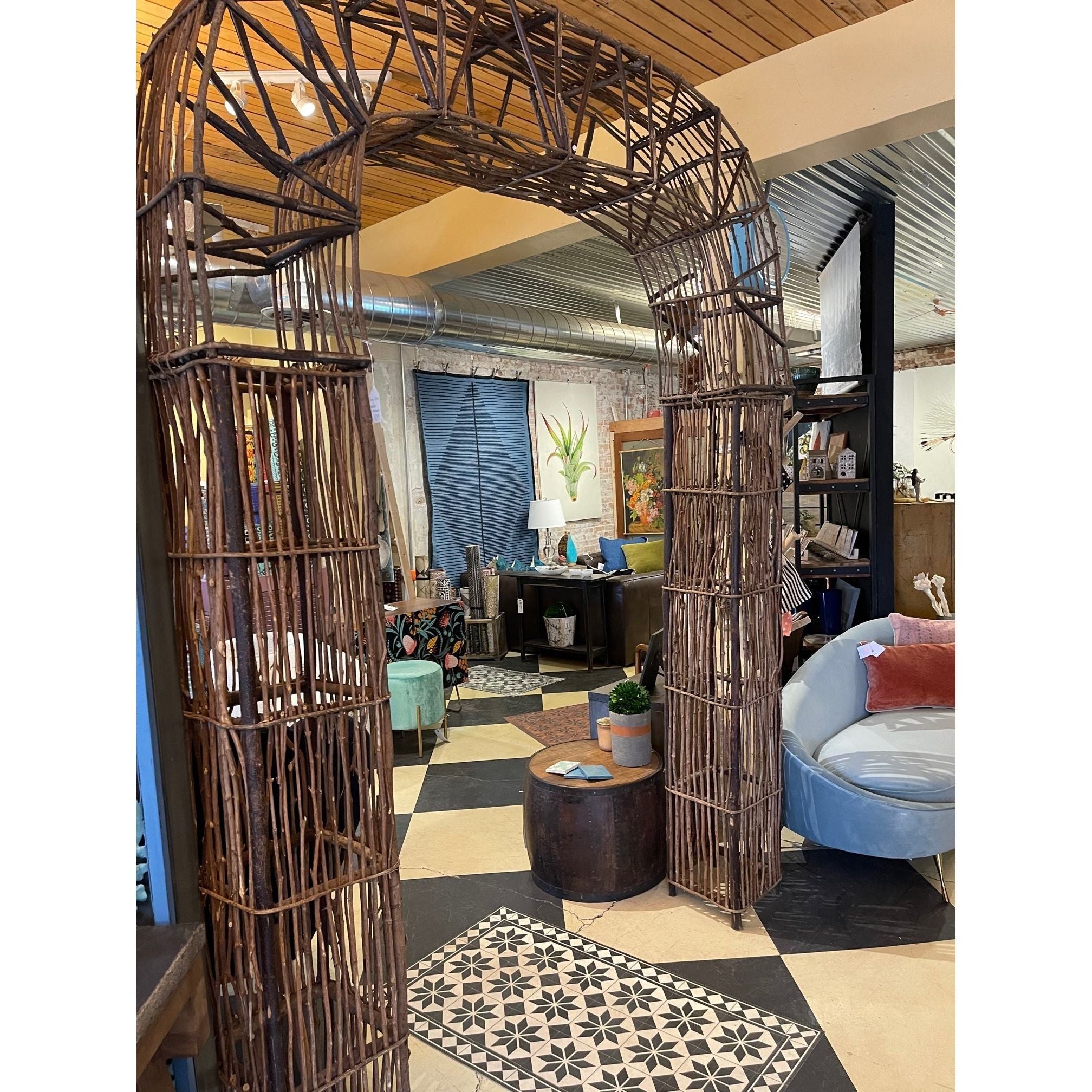 The Indoor/Outdoor Garden Arch, crafted from natural wood twigs, stands in a cozy room with checkered tiles, a round wooden table with a plant, a curved gray sofa with pillows, wall art, and customizable decor.