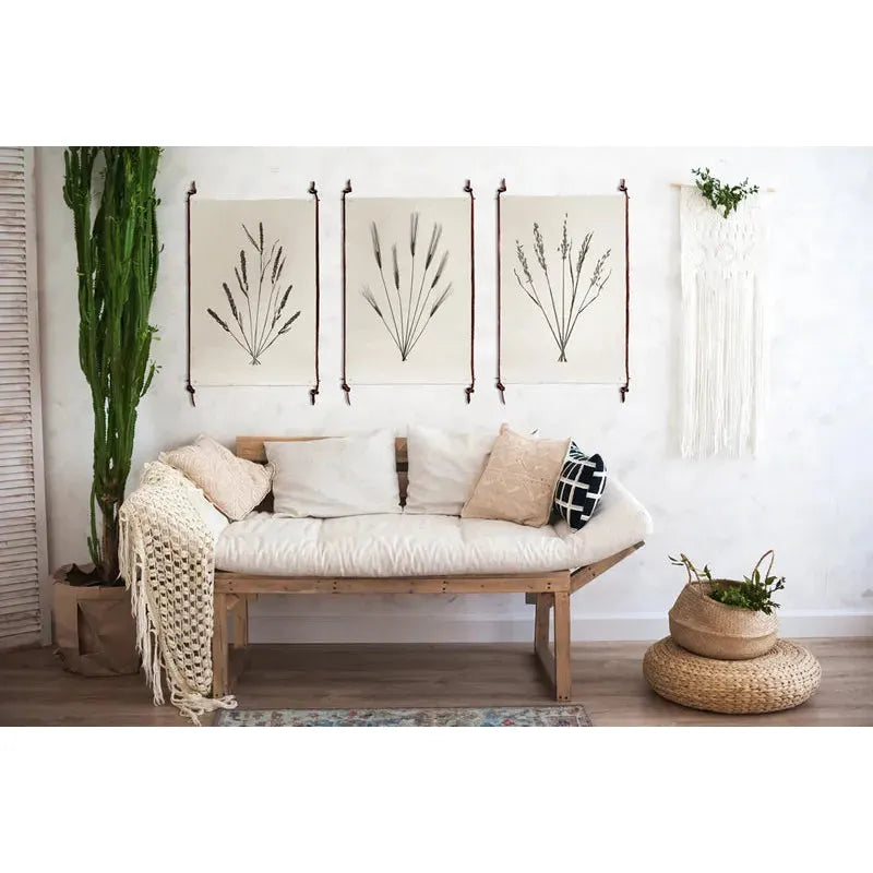 A cozy living space featuring a wooden bench sofa adorned with white and beige cushions and a knitted throw. Above the sofa, botanical prints are elegantly displayed using frameless leather/steel hangers that ensure non-damaging support. A tall cactus stands to the left, while to the right, a wicker basket containing a plant adds a natural touch on the floor.