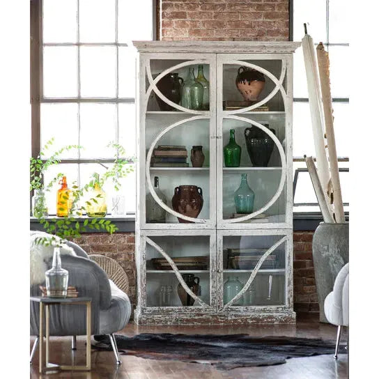 The Sandi Hutch, a vintage white cabinet made of pine wood with glass doors and geometric designs, displays eclectic vases and pottery. In a rustic room featuring large windows, exposed brick walls, a gray armchair, and lush plants, natural light enhances the cozy artistic vibe.