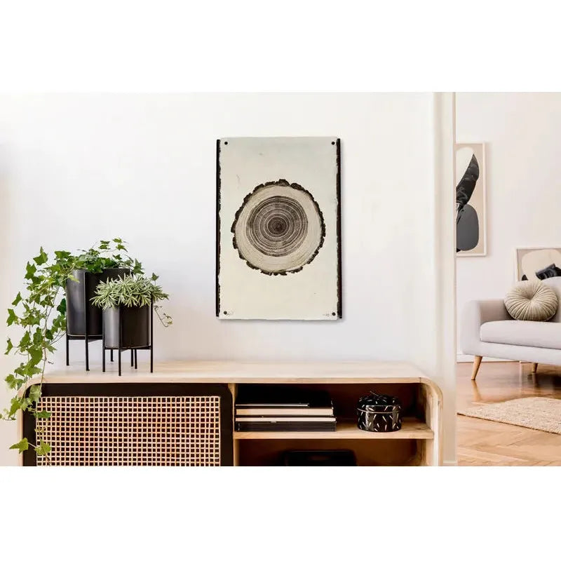 A minimalist interior with a wooden side table against a plain white wall. On the table are black and white plants in black pots. Above the table, showcasing a tree ring pattern, a framed artwork hangs securely from Frameless Wood/Steel Hanger, Straight. In the adjacent room, a light grey sofa with green and grey cushions is partially visible.