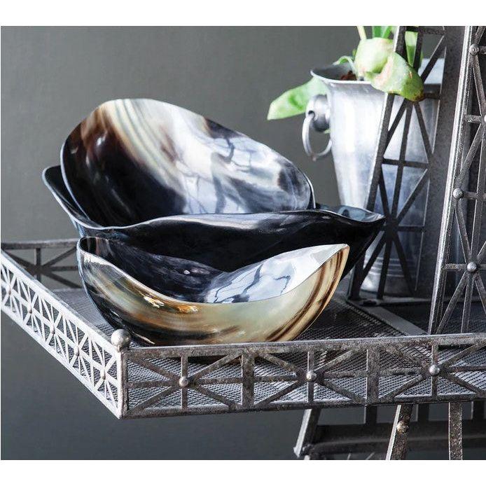 Two elegantly stacked, uniquely shaped Cow Horn Bowls feature swirling patterns in shades of black, brown, and ivory. They rest on a decorative metal shelf with lattice accents next to a metal pot holding a green plant, adding natural accents against the muted dark background.