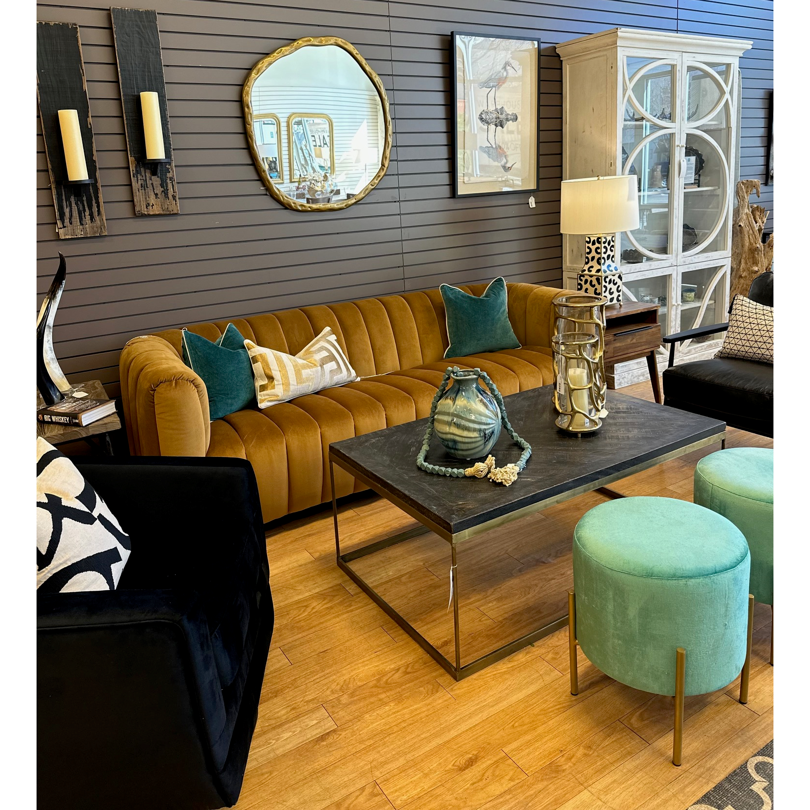 A modern living room setup features the Donna Upholstered Sofa in mustard with teal and brown pillows. A black coffee table displays decorative items, accompanied by two teal stools, a circular mirror, wall sconces, artwork, a sculptural dark armchair, and a glass-front cabinet.
