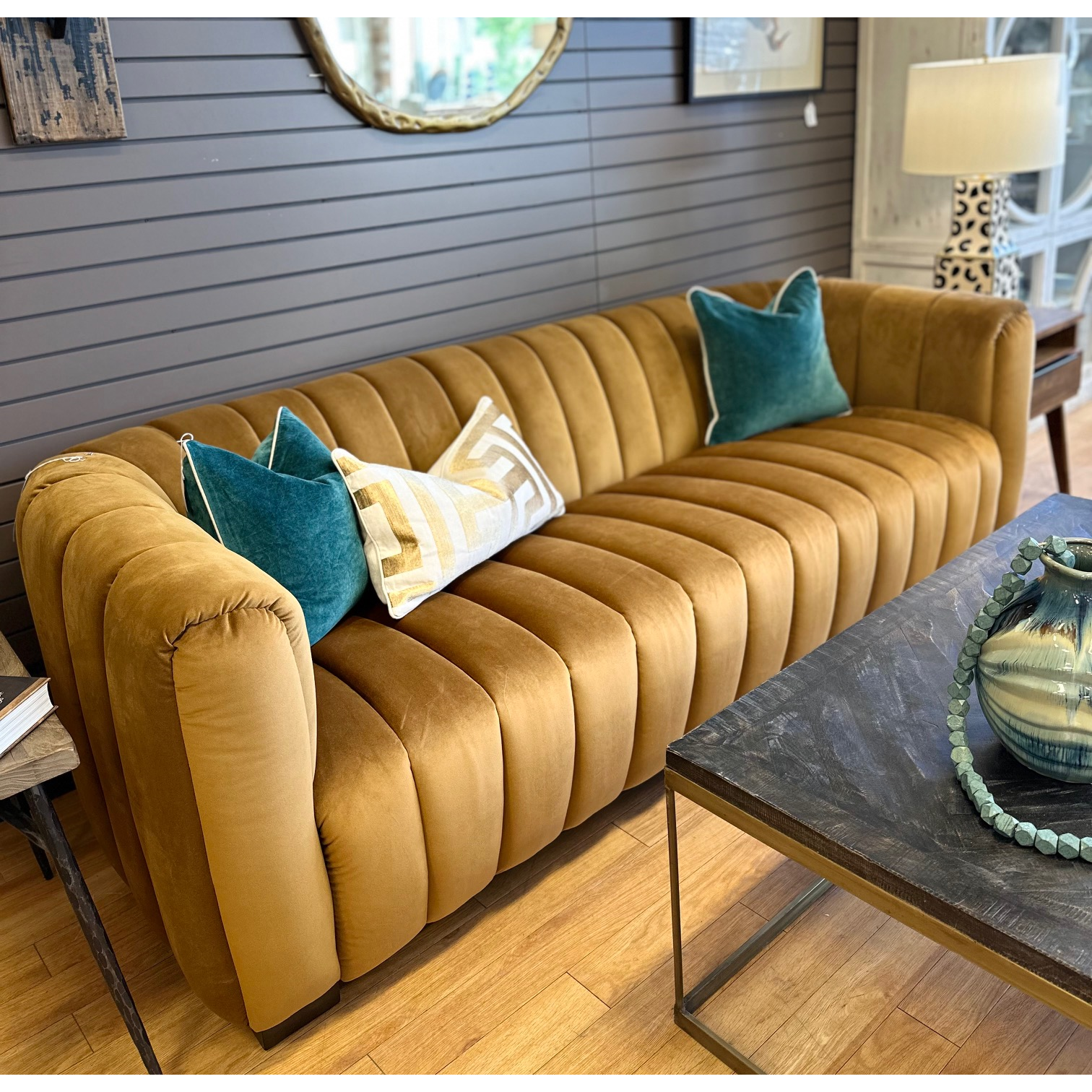 The Donna Upholstered Sofa, a plush mustard velvet piece with sculptural curves and vertical channel tufting, features two teal throw pillows and a cream geometric pillow. It pairs perfectly with the dark-paneled wall, round mirror above, wooden coffee table, and ceramic vase.