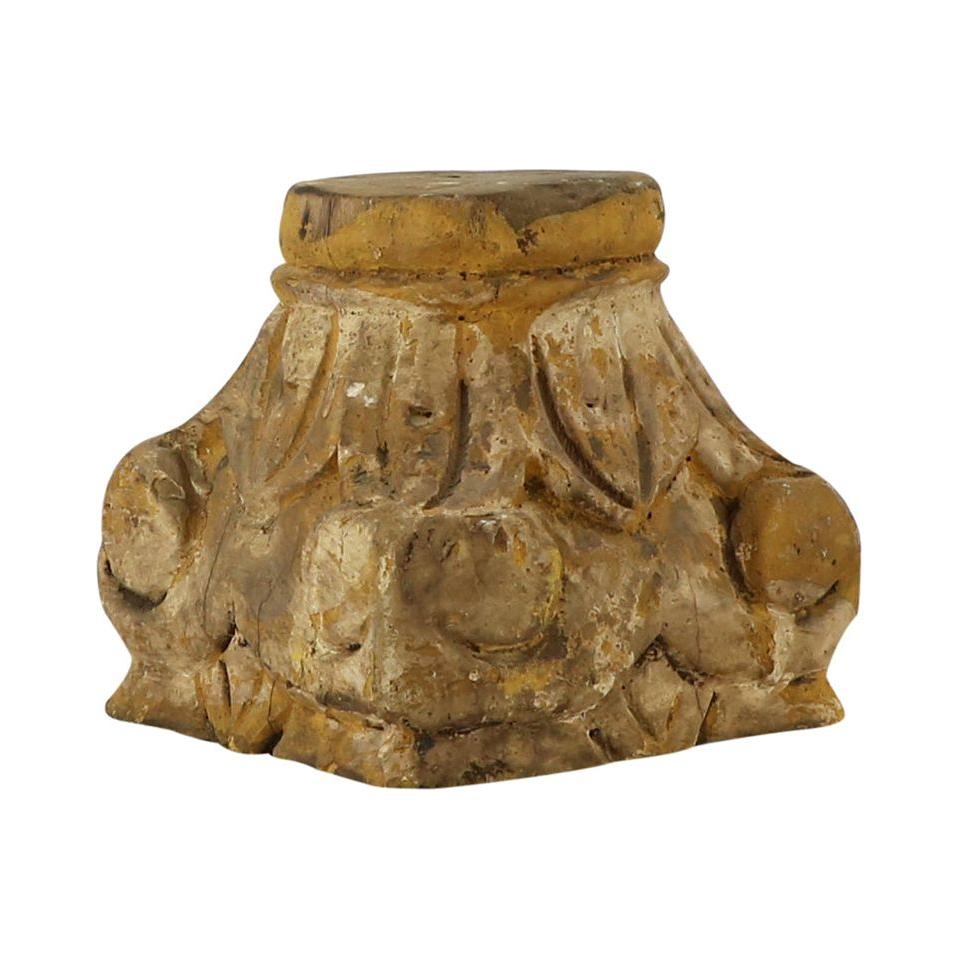 The Carved Pillar Base in natural wood showcases a weathered stone capital with hand-carved floral and leaf motifs. Its rustic brown patina, worn surface, and rough edges evoke antiquity. The capital sits atop the intricately designed, wide pillar base with a flat top.